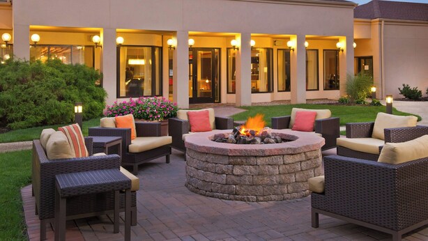 An outdoor patio with wicker furniture, a stone fire pit, and a building with illuminated windows and doors. - Courtyard by Marriott Mt. Laurel (Mount Laurel)