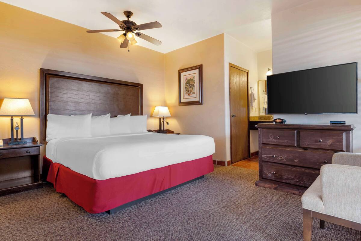 Habitación de hotel con cama, dos lámparas de noche, ventilador de techo, cómoda, televisor y un cuadro en la pared.