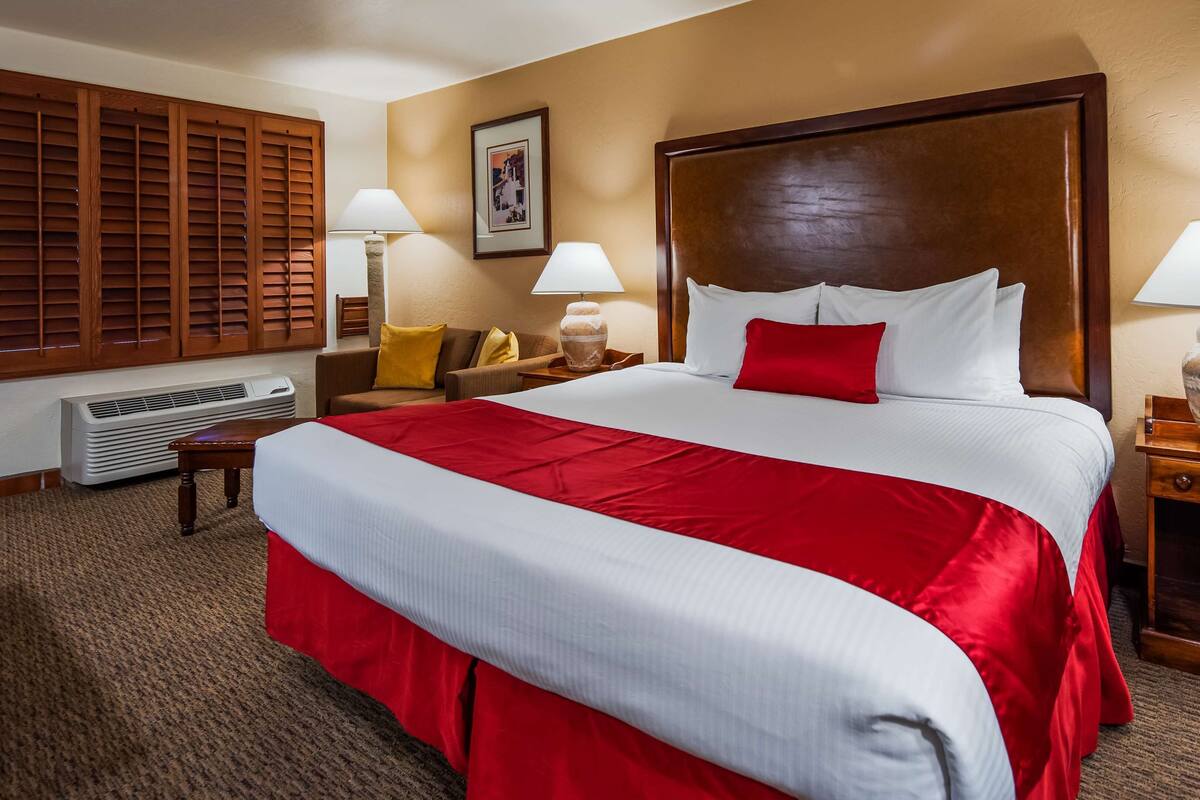 Habitación de hotel con una cama grande, dos mesitas de noche, una silla, un cuadro en la pared y una ventana con persianas.