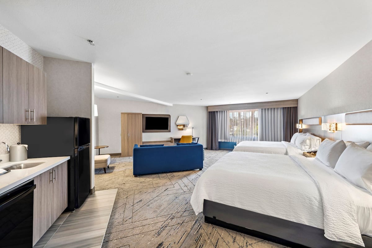 A modern hotel room with a kitchenette, a large bed, a sofa, and a TV.