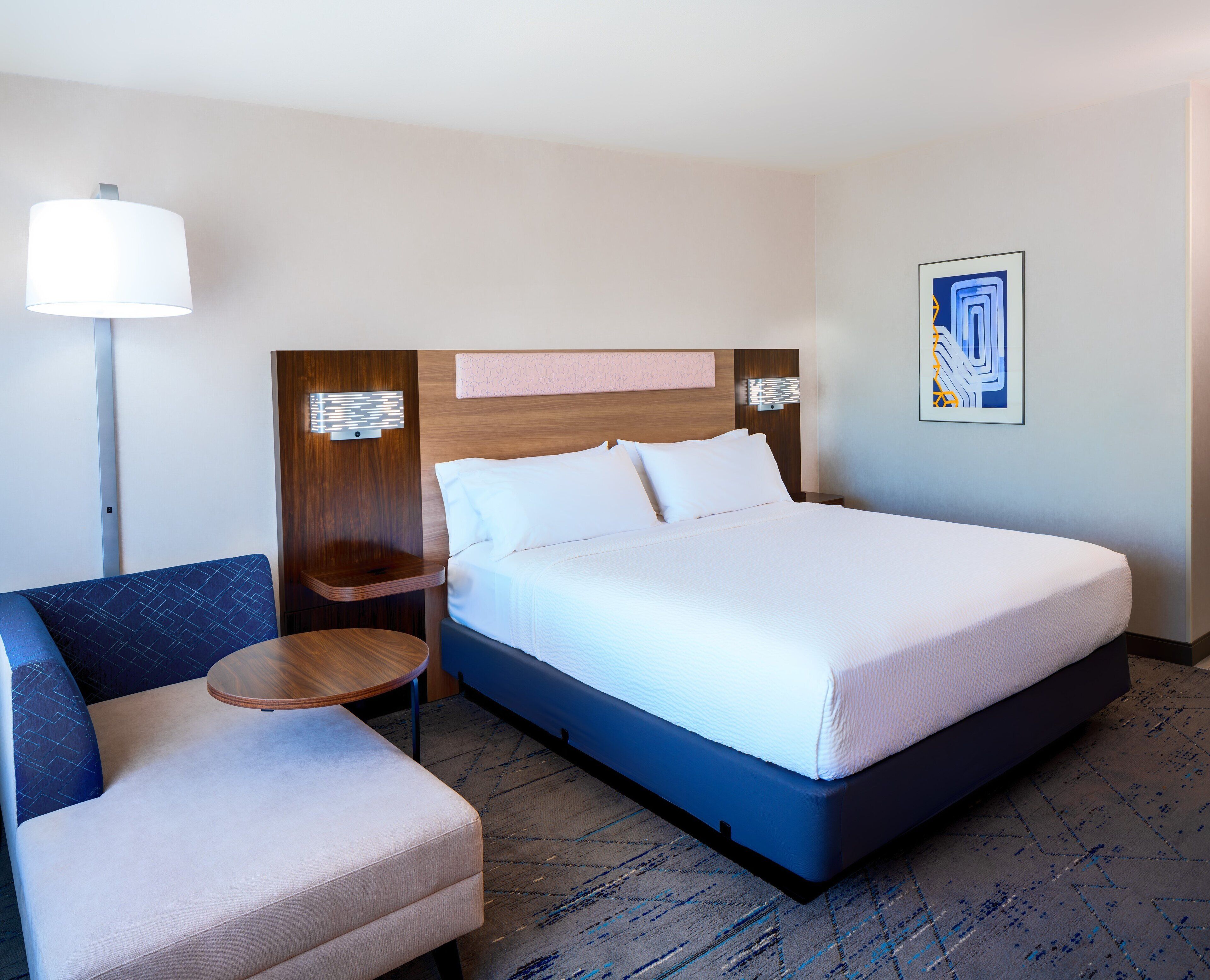 A hotel room with a bed, a chair, a bedside table, a lamp, and a wall art piece.