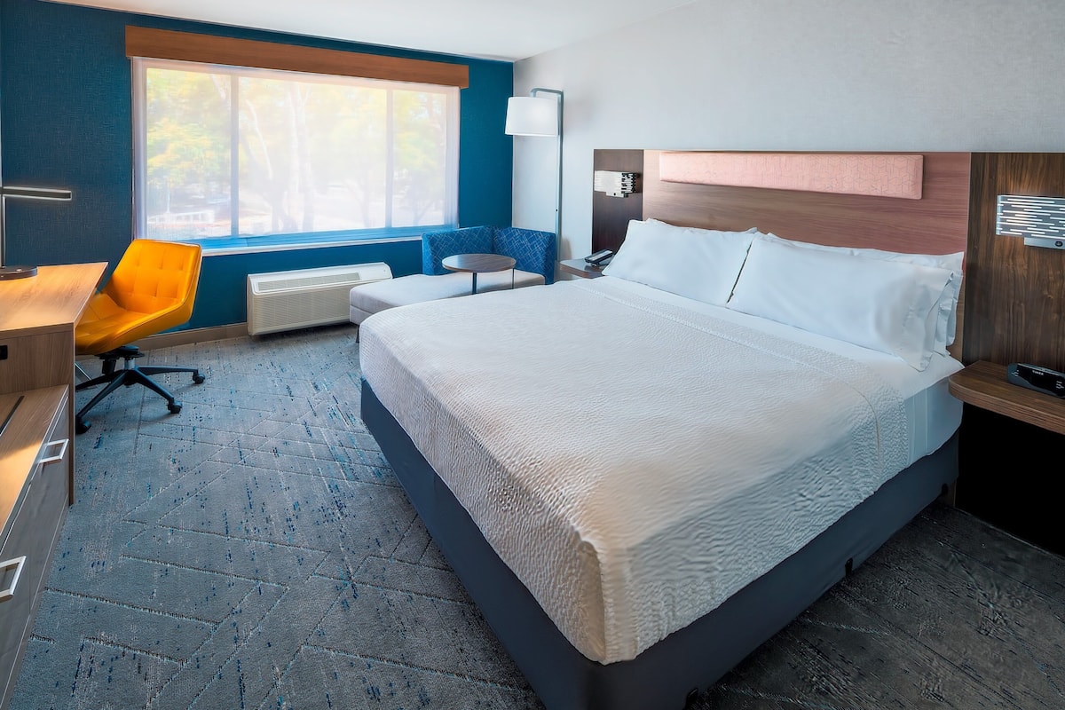 A modern hotel room with a large bed, a desk, a chair, and a window with a view of trees.