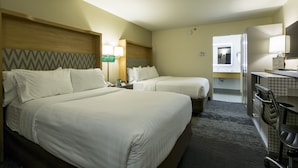 Une chambre d’hôtel avec deux lits, un bureau, une chaise, un miroir et une salle de bain visible à travers une porte ouverte.