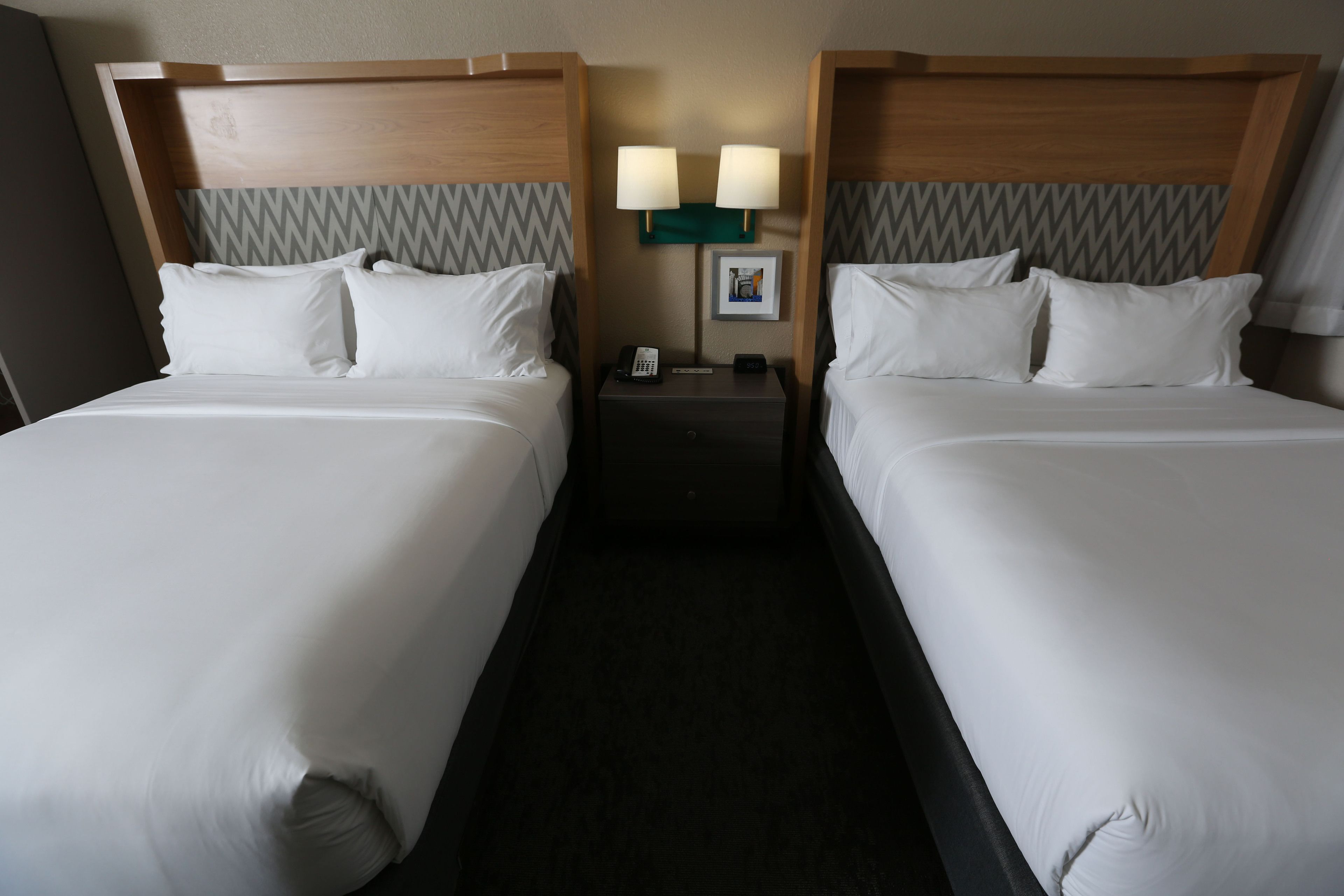 A hotel room with two beds, each with white linens and pillows, a nightstand with a phone and lamp, and a wall-mounted picture.