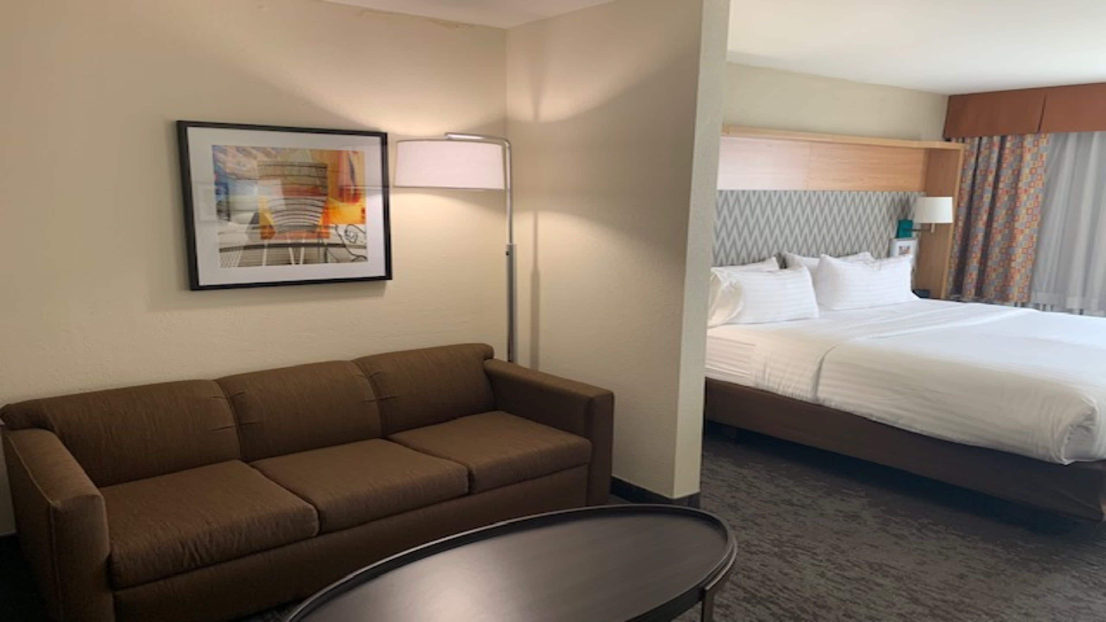 A hotel room with a brown sofa, a round table, a bed with white bedding, and a framed picture on the wall.