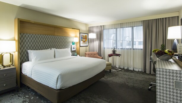 Une chambre d’hôtel avec un grand lit, une chaise, un bureau avec une lampe, un panier de fruits et une fenêtre avec des rideaux.