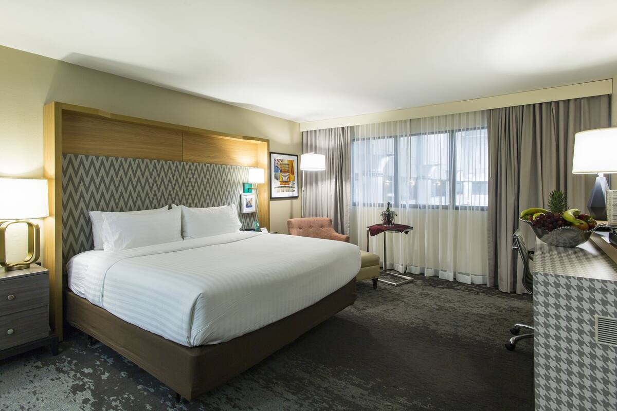 A hotel room with a large bed, a chair, a desk with a lamp, a fruit bowl, and a window with curtains.
