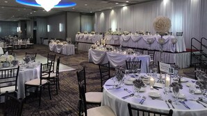 Une salle de banquet avec des tables rondes dressées pour un événement formel, agrémentée de nappes blanches, de compositions florales et d’un éclairage à la bougie.