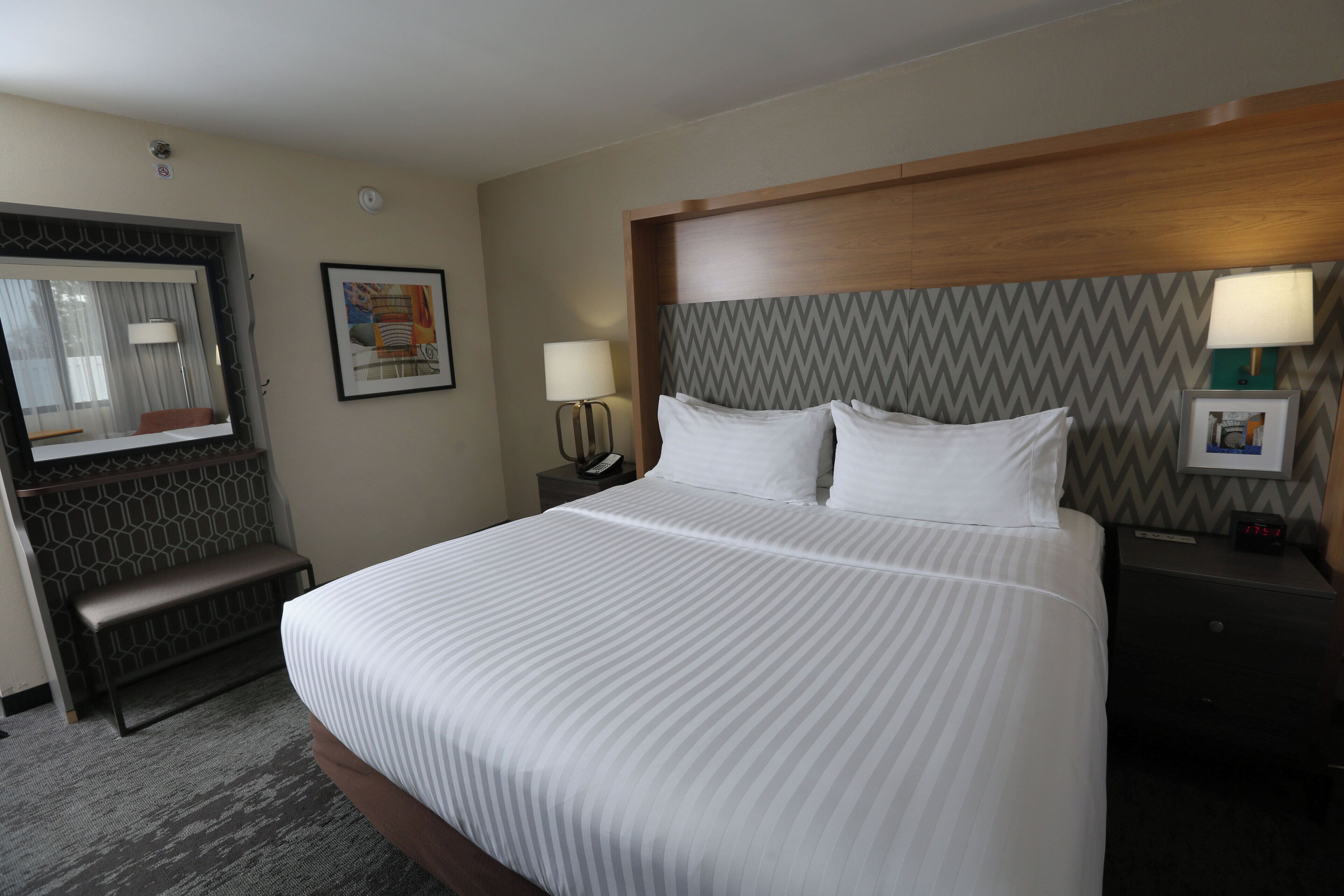 A hotel room with a large bed, a desk, a chair, a lamp, a phone, and a clock.