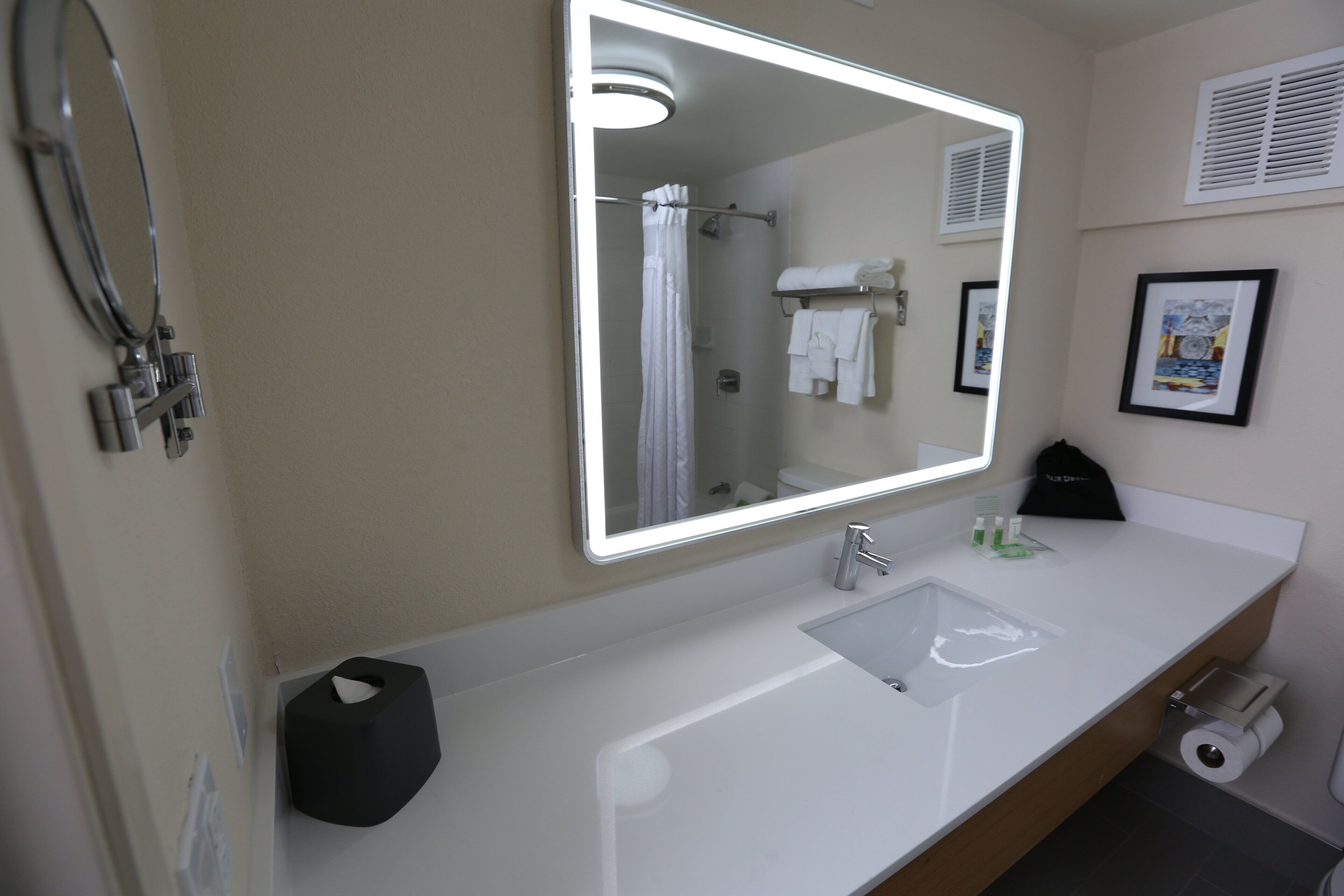 Une salle de bain d’hôtel avec un grand miroir, un lavabo, un porte-serviettes, un rideau de douche et un porte-rouleau de papier toilette.