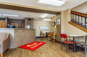 A hotel lobby with a reception desk, a dining area with chairs and a table, a clock on the wall, and a staircase.