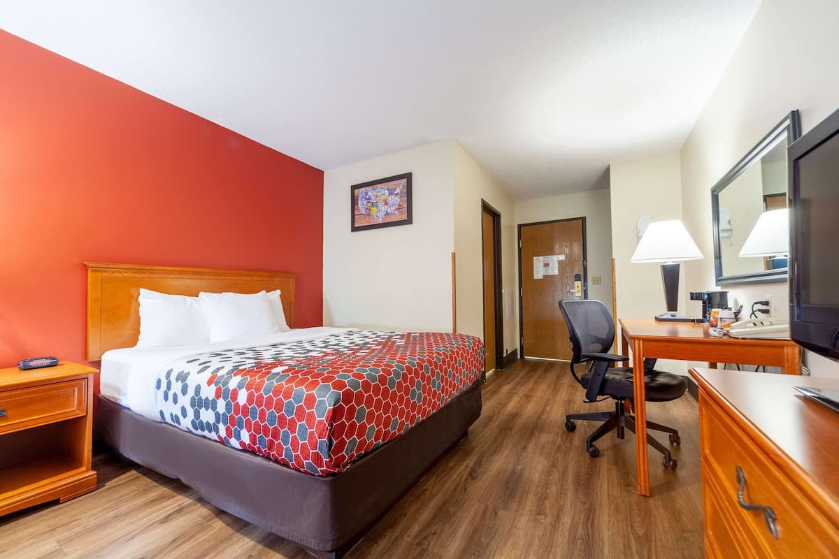 A hotel room with a bed, a desk, a chair, a nightstand, a lamp, and a television.