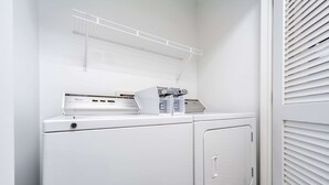 Un área de lavandería con lavadora y secadora, un estante sobre la lavadora, y una puerta con persianas.