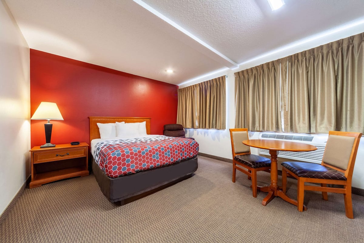 A hotel room with a bed, bedside table, lamp, and a dining area with a table and chairs.
