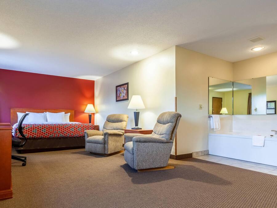 A hotel room with a bed, two armchairs, a desk, a lamp, a large mirror, and a bathroom with a sink and towel.