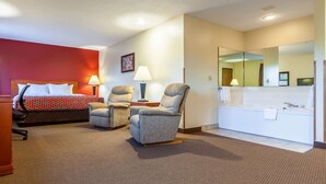 Habitación de hotel con cama, dos sillones, escritorio, lámpara, espejo grande y baño con lavamanos y toalla.