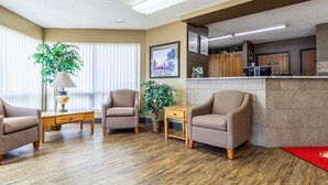 Un vestíbulo de hotel con paredes color beige, suelo de madera y un mostrador de recepción. Hay tres sillones color beige, una mesita de madera y dos plantas en macetas.