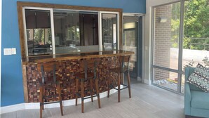 A bar area with a wooden counter and three stools, a large window with a view of greenery, and a blue wall. - Wingate by Wyndham Columbia/Fort Jackson (Columbia)