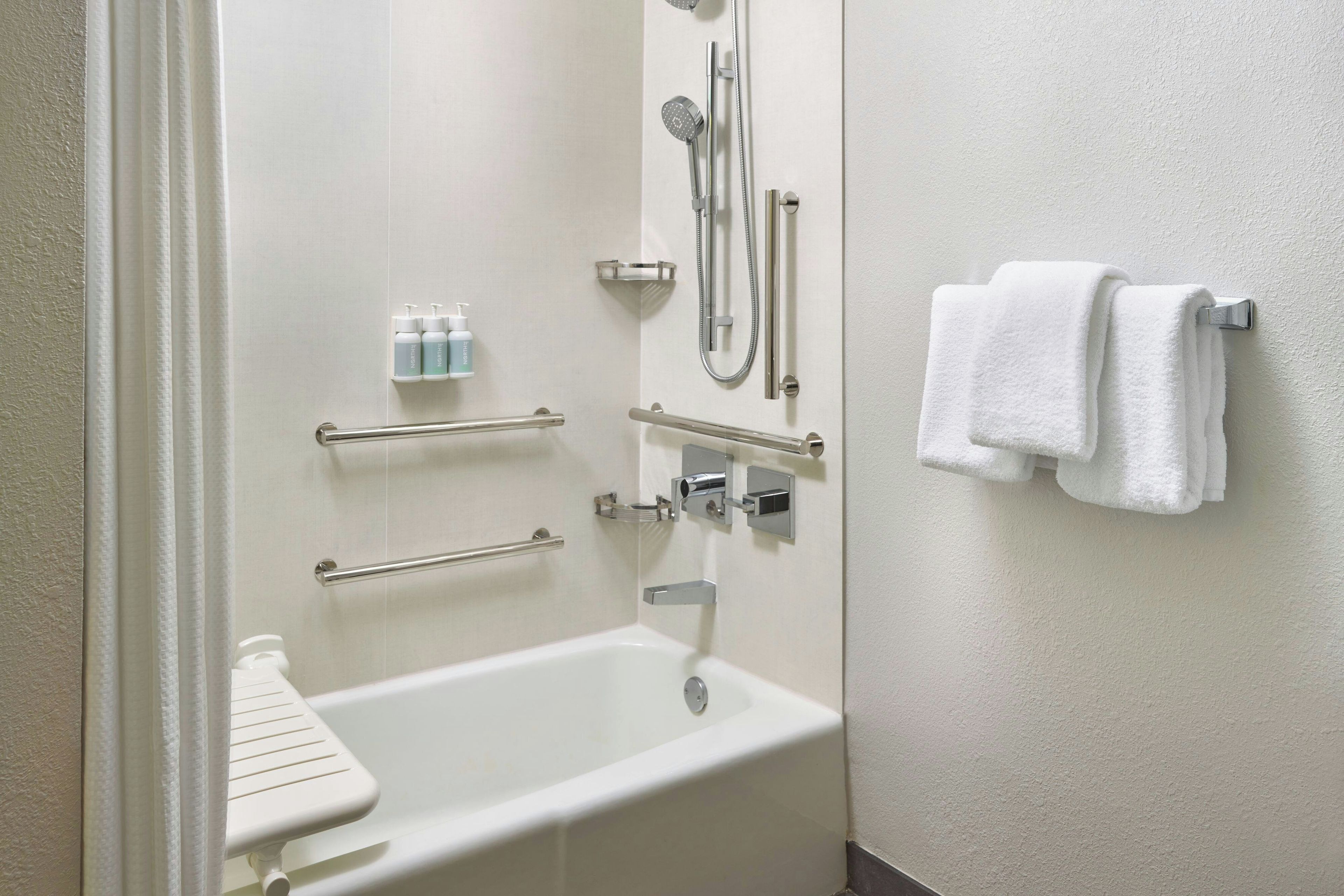 A bathroom with a bathtub, a shower, and handrails. There are two bottles of hand soap on the wall.