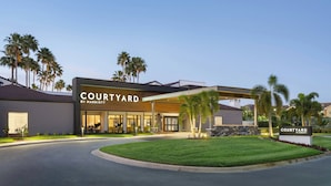 Eingang eines Courtyard by Marriott Hotels mit überdachtem Gang, Palmen und einem gepflegten Rasen.