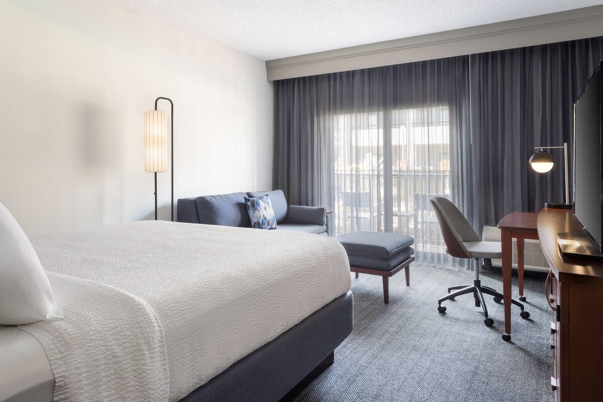 Une chambre d’hôtel avec un grand lit, un bureau avec une chaise, une lampe sur pied et une vue sur l’extérieur par une fenêtre munie de rideaux.