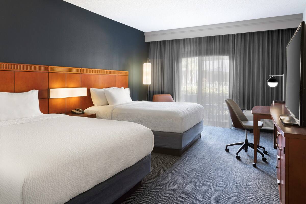 Une chambre d’hôtel avec deux lits, un bureau, une chaise et une grande fenêtre avec des rideaux.