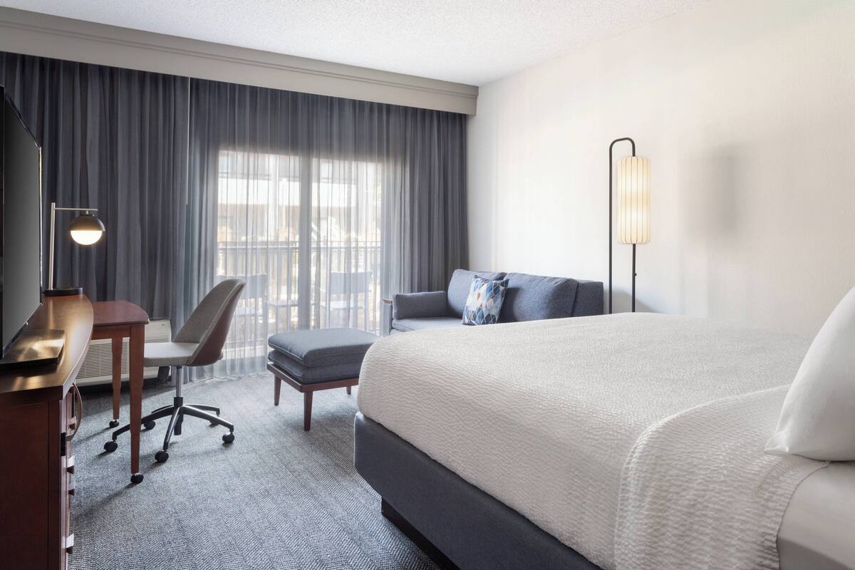 Une chambre d’hôtel avec un grand lit, un bureau avec une chaise, un lampadaire et une vue sur l’extérieur à travers des rideaux légers.