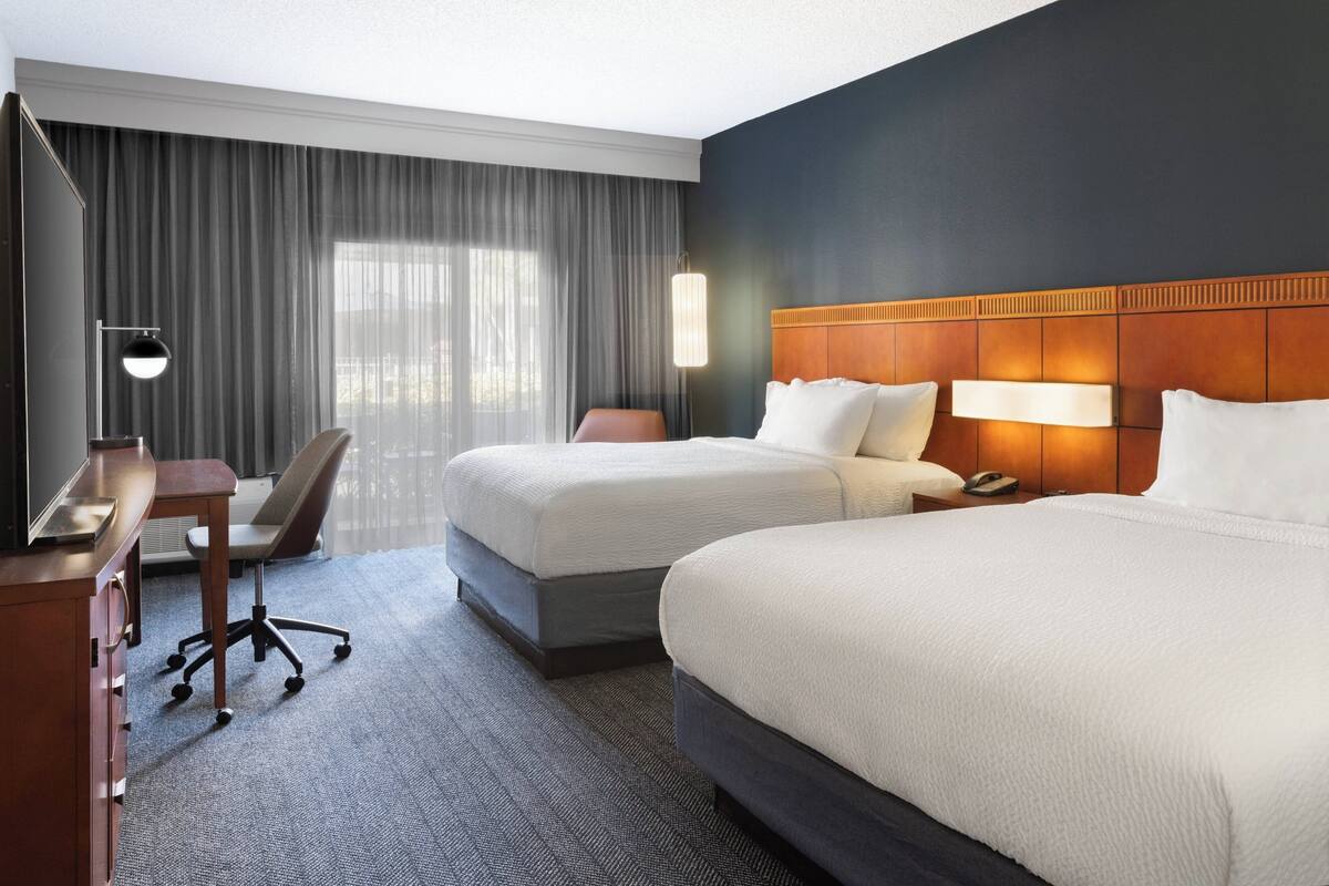 Une chambre d’hôtel avec deux lits, un bureau, une chaise et une grande fenêtre avec des rideaux.