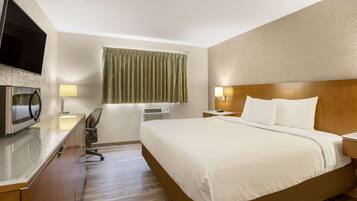 Una habitación de hotel con cama, escritorio con computadora, televisor de pantalla plana, lámpara y ventana con cortinas.
