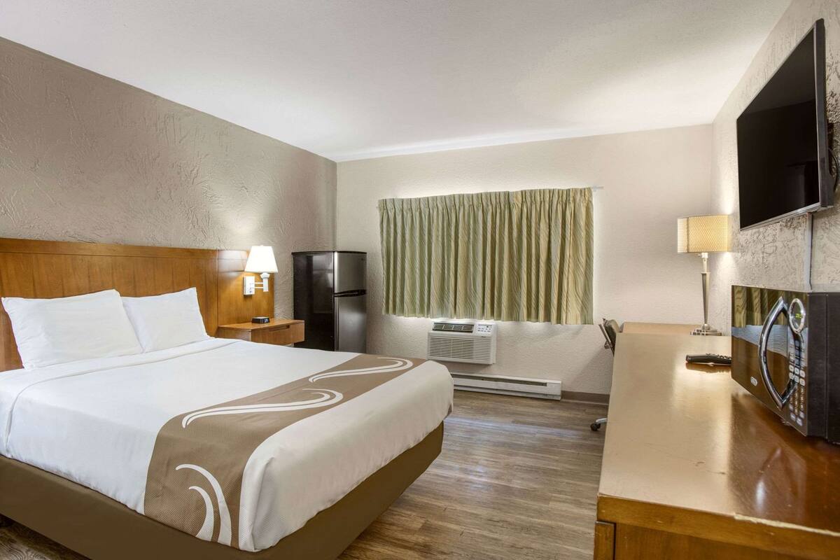 Una habitación de hotel con cama, escritorio, televisor, refrigerador y aire acondicionado.