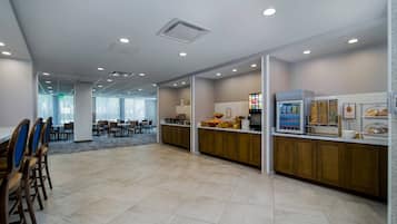 A spacious dining area with a long counter and multiple tables, a kitchen area with a refrigerator and food display, and a corridor leading to another room.
