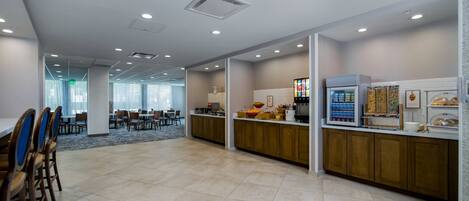 A spacious dining area with a long counter and multiple tables, a kitchen area with a refrigerator and food display, and a corridor leading to another room.