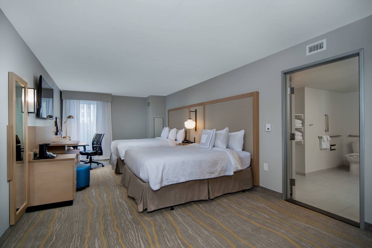 Habitación de hotel con dos camas, un escritorio, una silla y un baño visible a través de un vidrio.