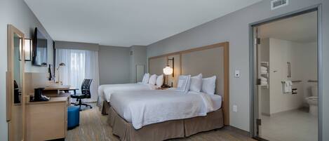 A hotel room with two beds, a desk, a chair, and a bathroom visible through a glass door.