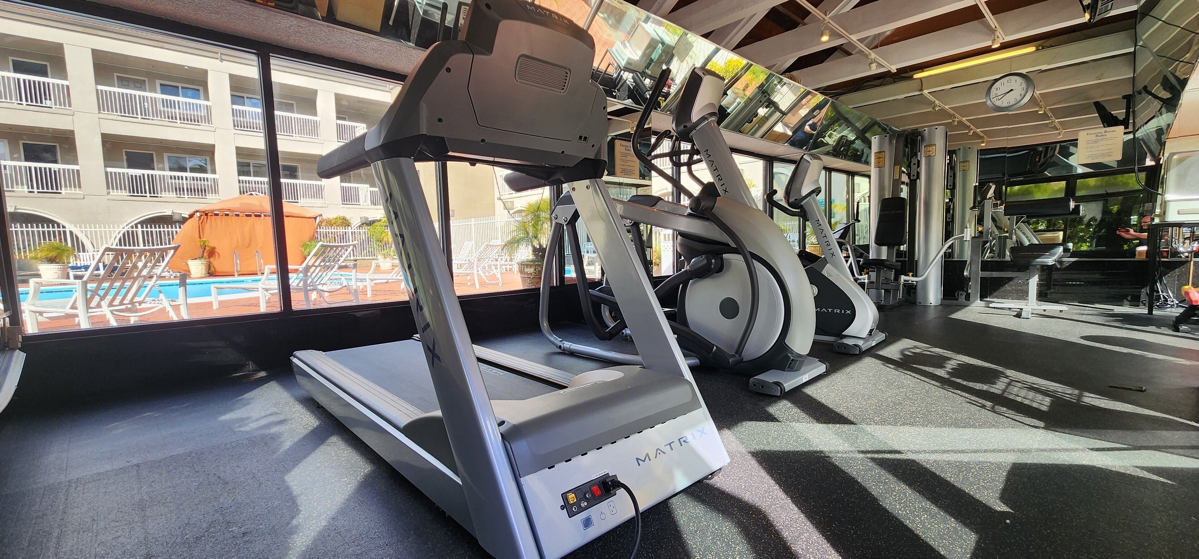 A gym with treadmills and exercise equipment, a pool visible through the window, and a building with balconies in the background.