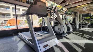 A gym with treadmills and exercise equipment, a pool visible through the window, and a building with balconies in the background.