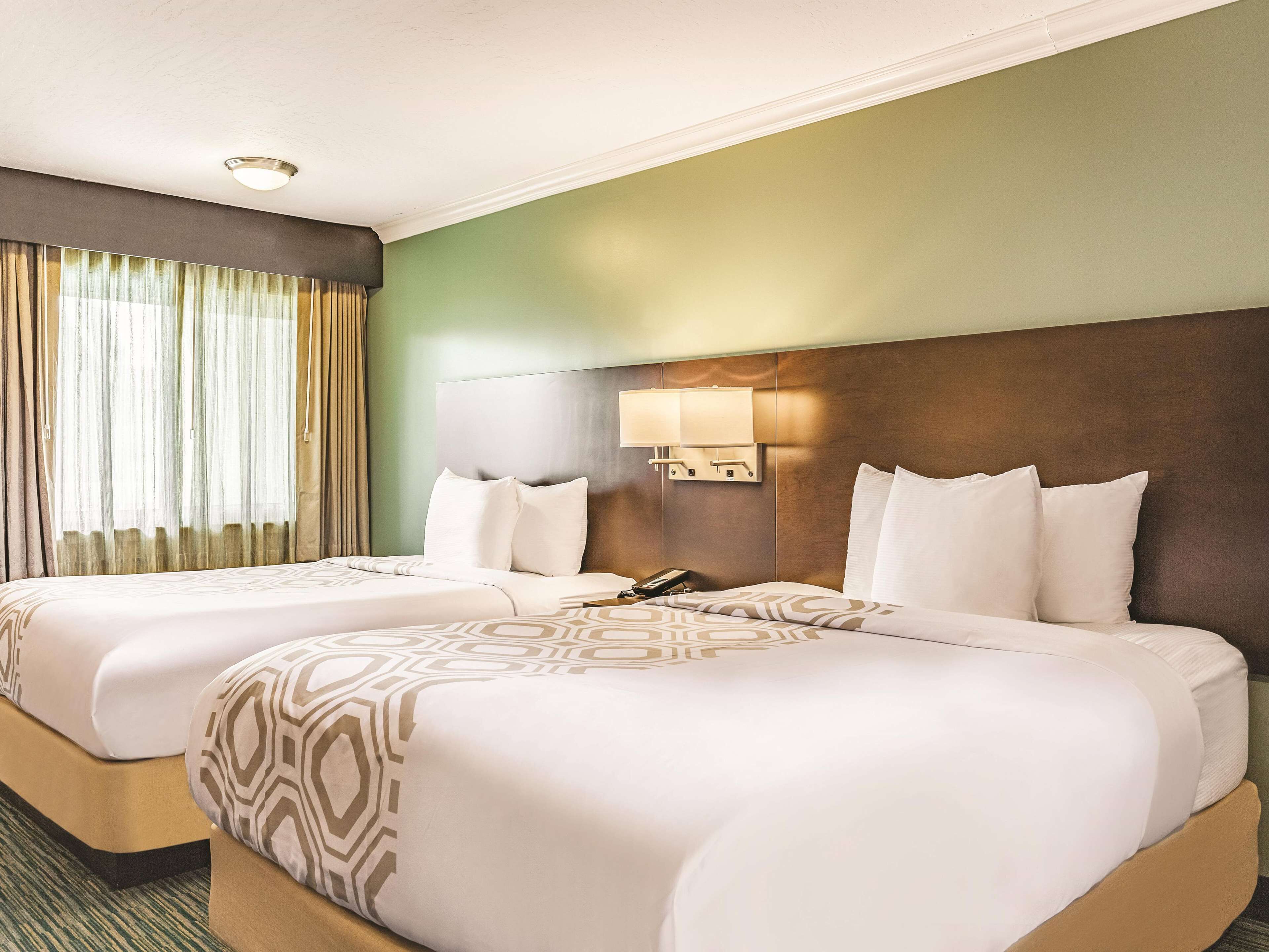 A hotel room with two beds, a headboard with a light fixture, a bedside table with a phone, and a window with curtains.