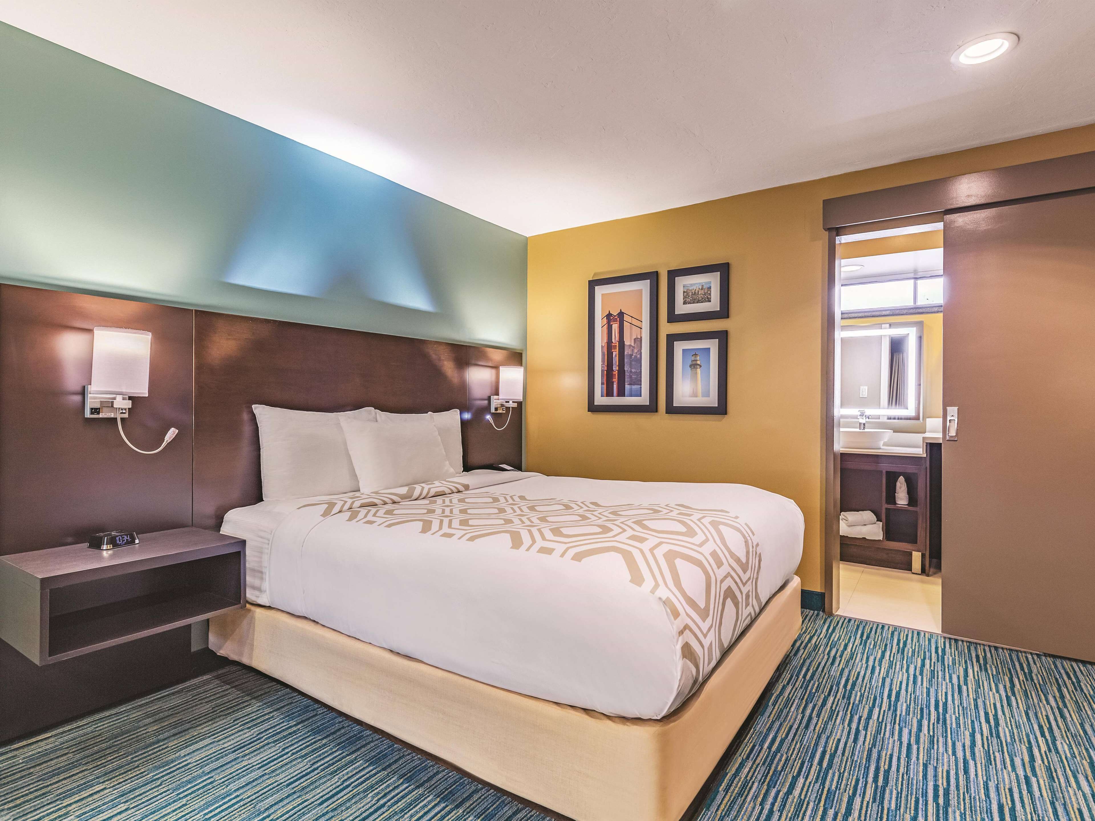 A hotel room with a large bed, bedside tables, a nightstand, a wall-mounted lamp, and a bathroom visible through an open door.