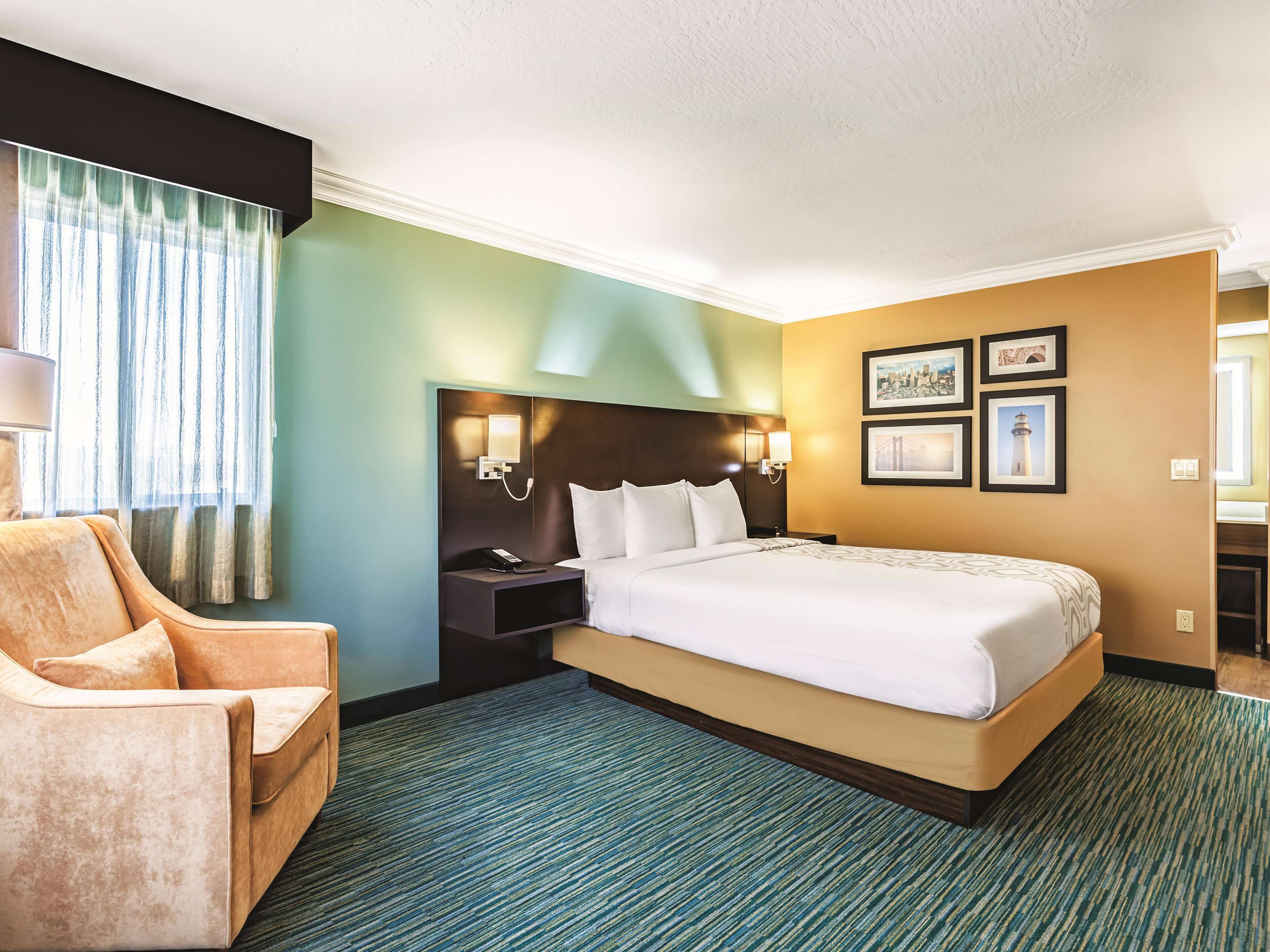 A hotel room with a large bed, a nightstand, a lamp, a chair, and framed pictures on the wall.
