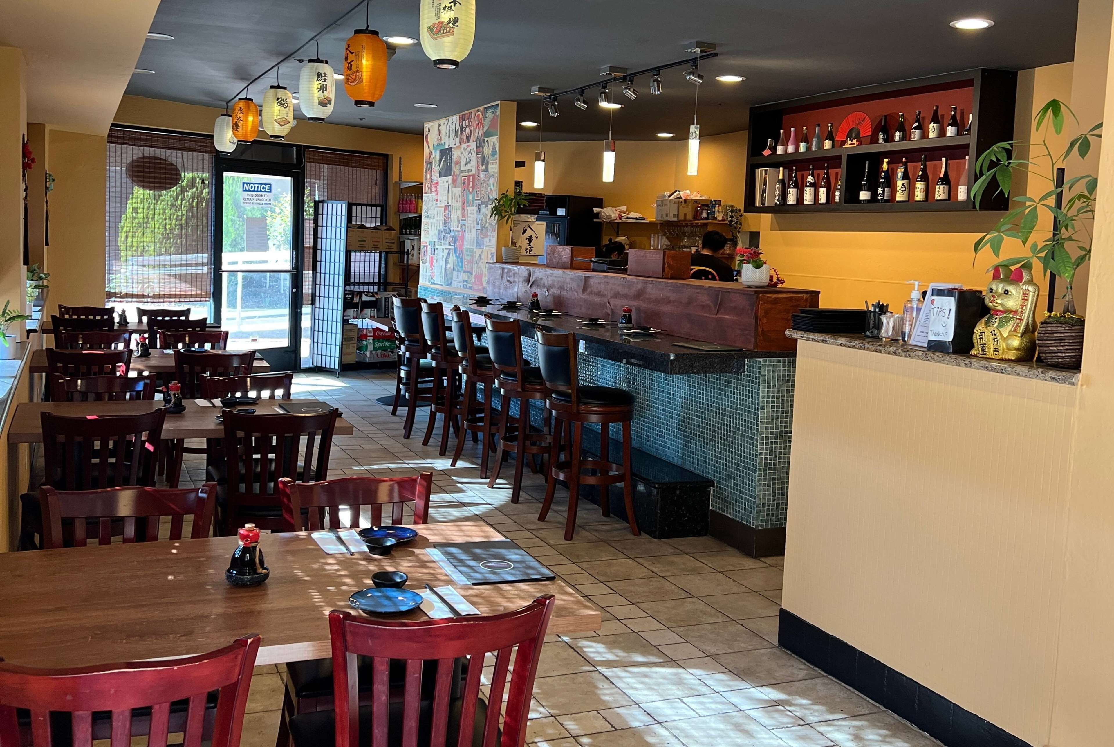A restaurant interior with wooden tables and chairs, a bar area, and shelves with bottles.