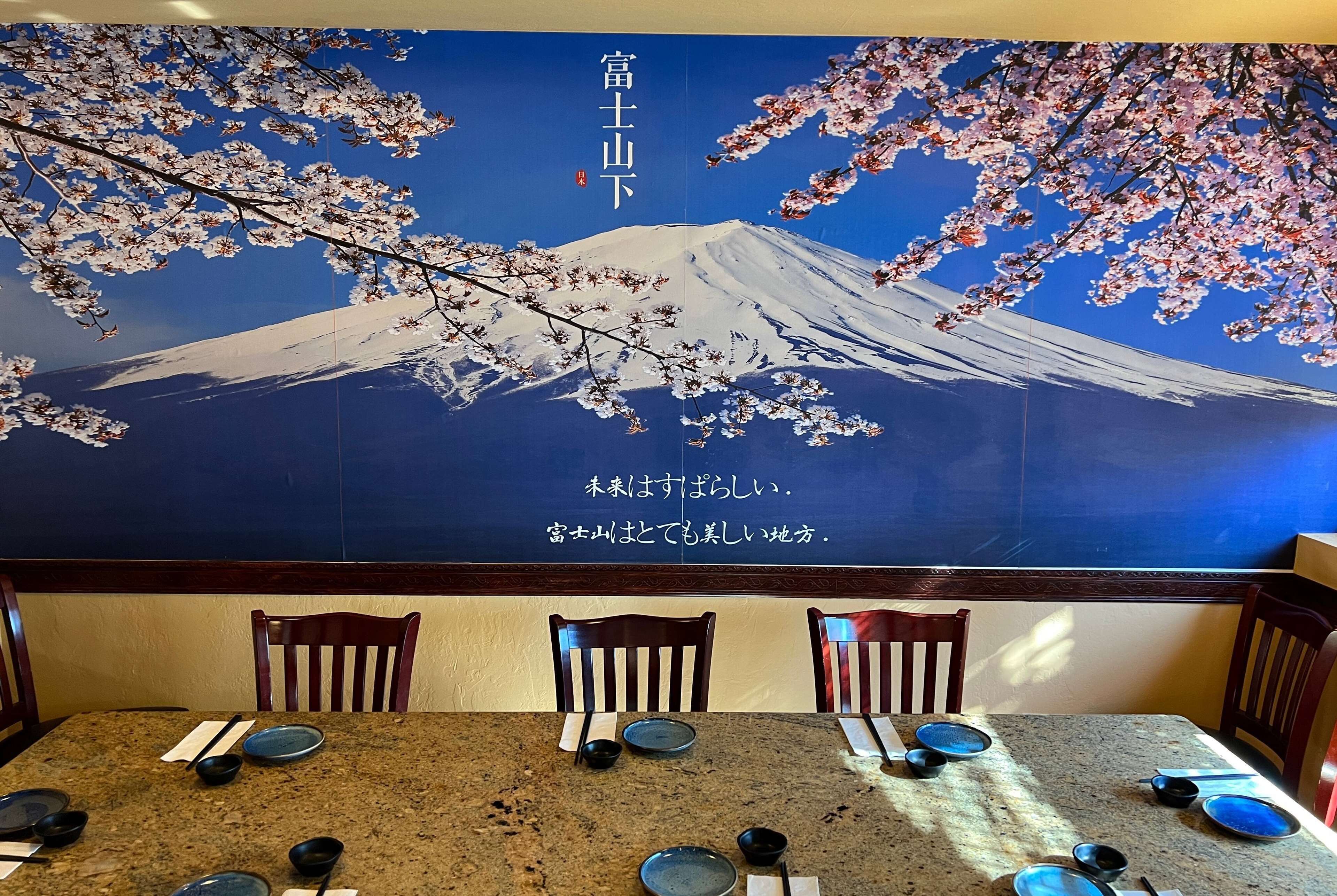 Un ristorante con un grande murale raffigurante il Monte Fuji e fiori di ciliegio. Sono apparecchiate tavole con piatti e ciotole.