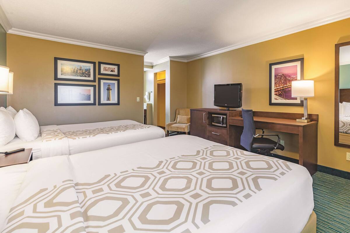 Habitación de hotel con dos camas, un escritorio con televisor, una silla y cuadros en la pared.
