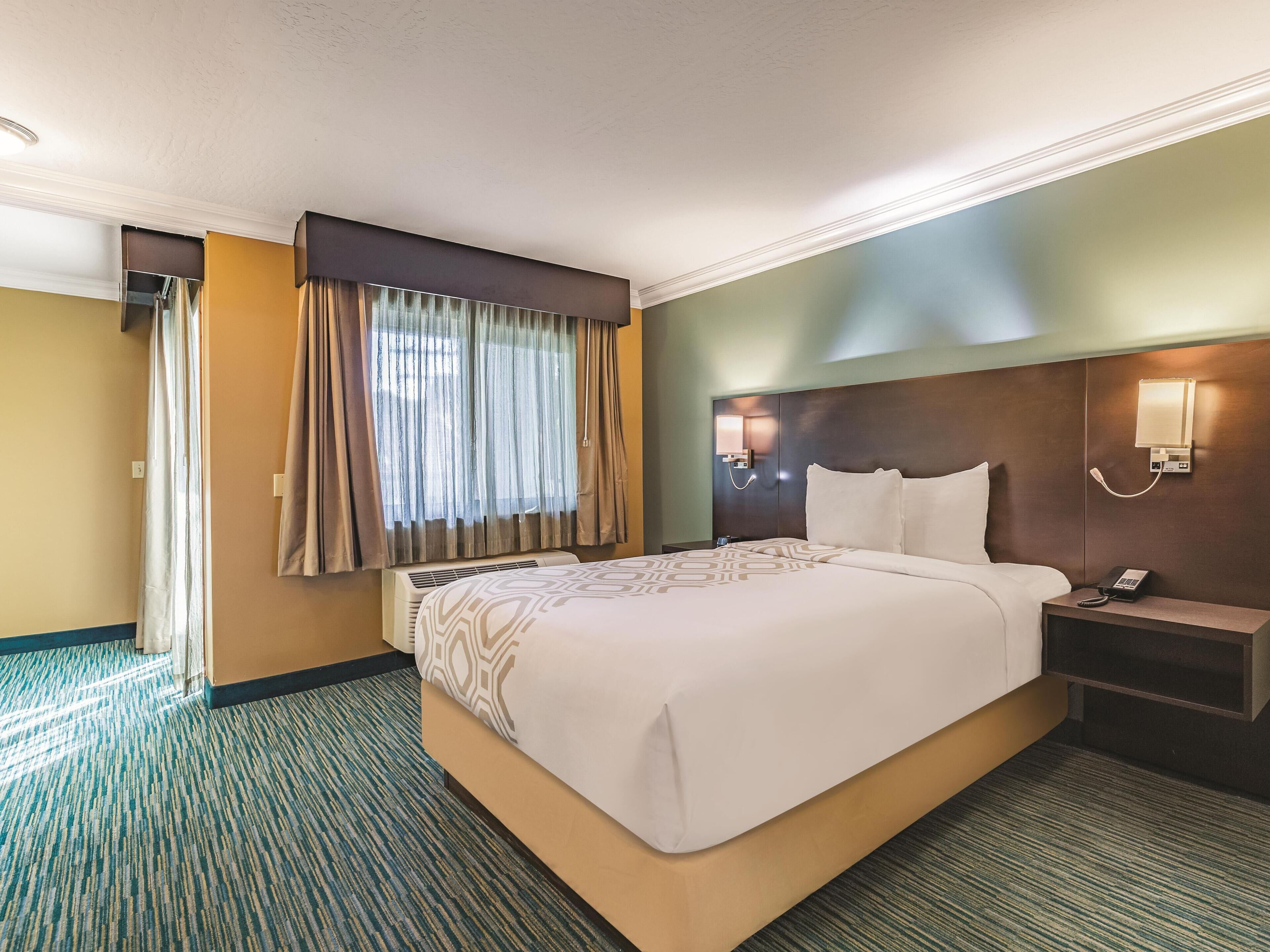 A hotel room with a large bed, a bedside table with a phone, a wall-mounted light, and a window with curtains.