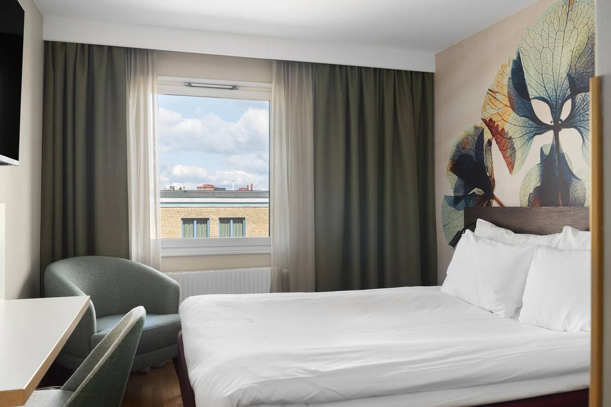 A hotel room with a bed, a desk, a chair, a window with curtains, and a wall painting.