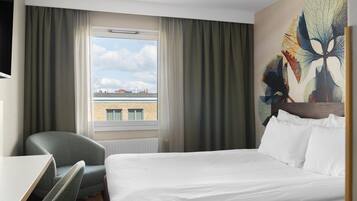 Habitación de hotel con cama, escritorio, silla, ventana con cortinas y un cuadro en la pared.
