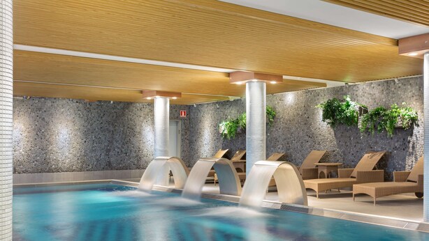 Une piscine intérieure moderne, dotée d’une eau claire, de chaises longues et d’un mur en pierre agrémenté de verdure.