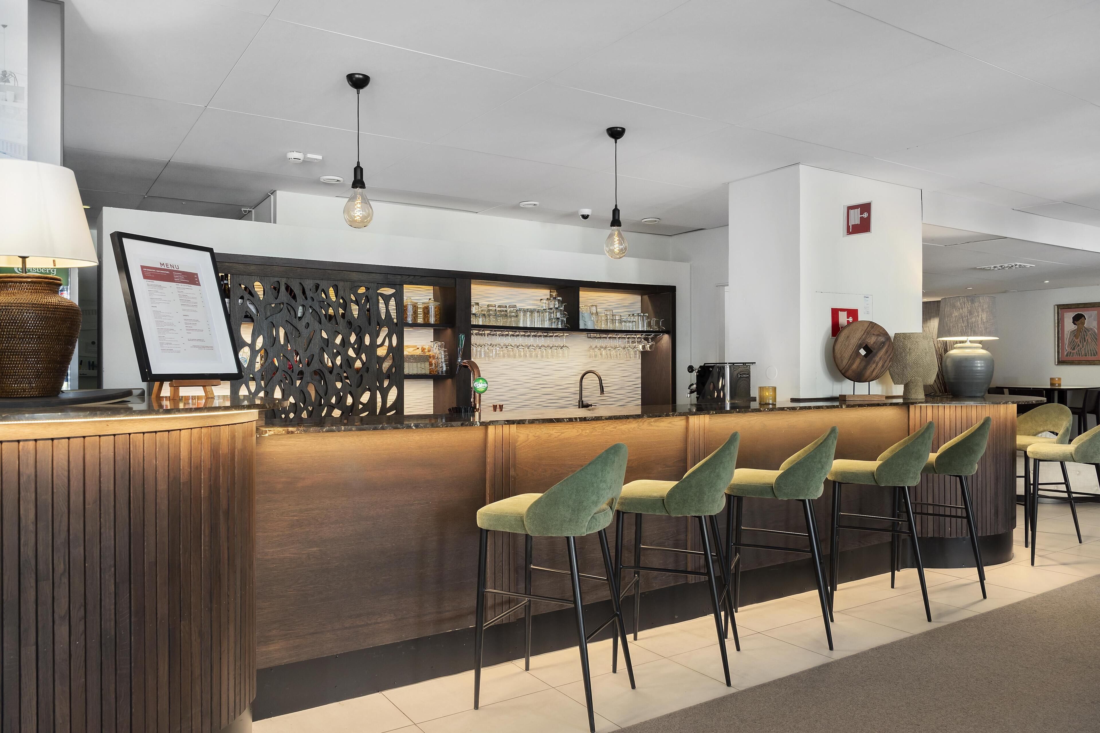 A bar area with wooden counter, green bar stools, hanging pendant lights, and a menu board.
