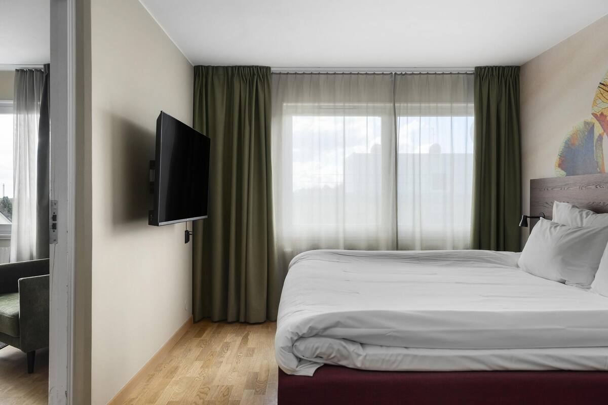 A hotel room with a bed, a television mounted on the wall, a chair, and a window with curtains.