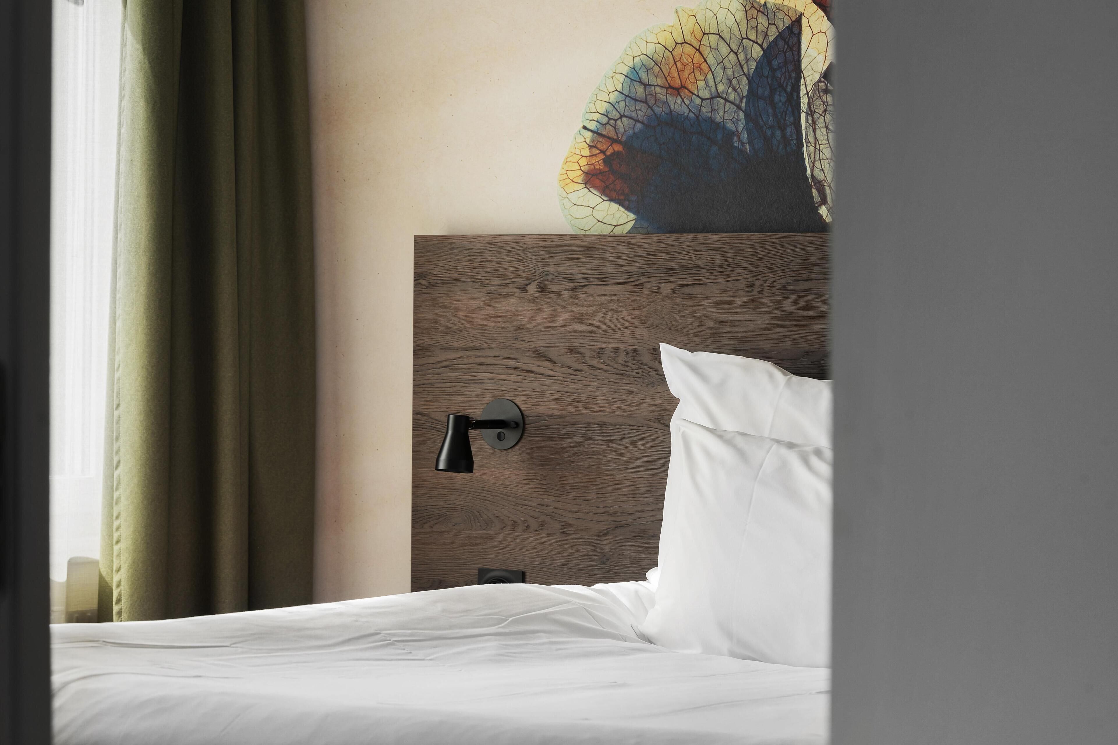 Habitación de hotel con cabecera de madera, lámpara de noche, almohada blanca y una obra de arte en la pared.