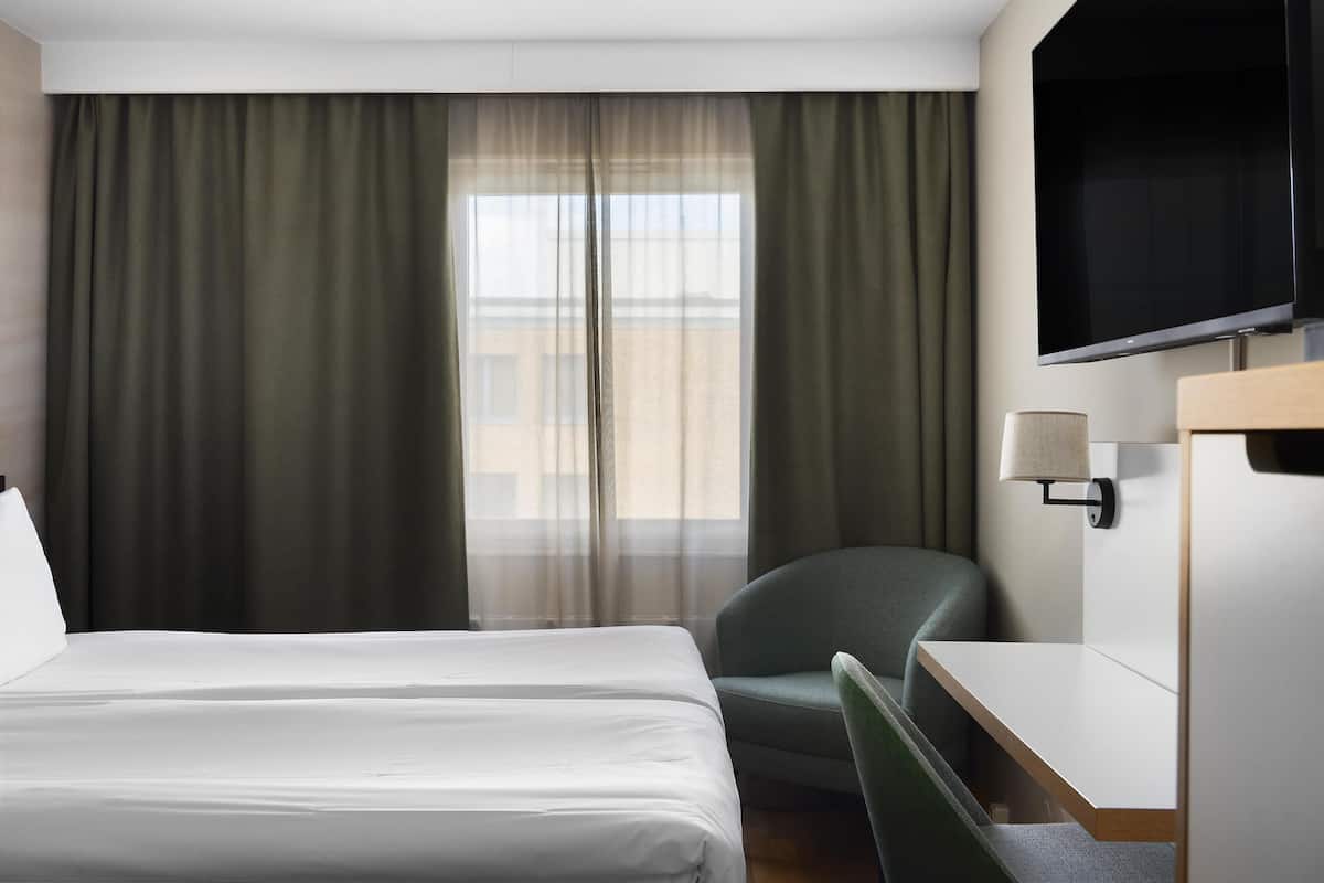 A hotel room with a bed, a chair, a desk, a TV, and a window with curtains.